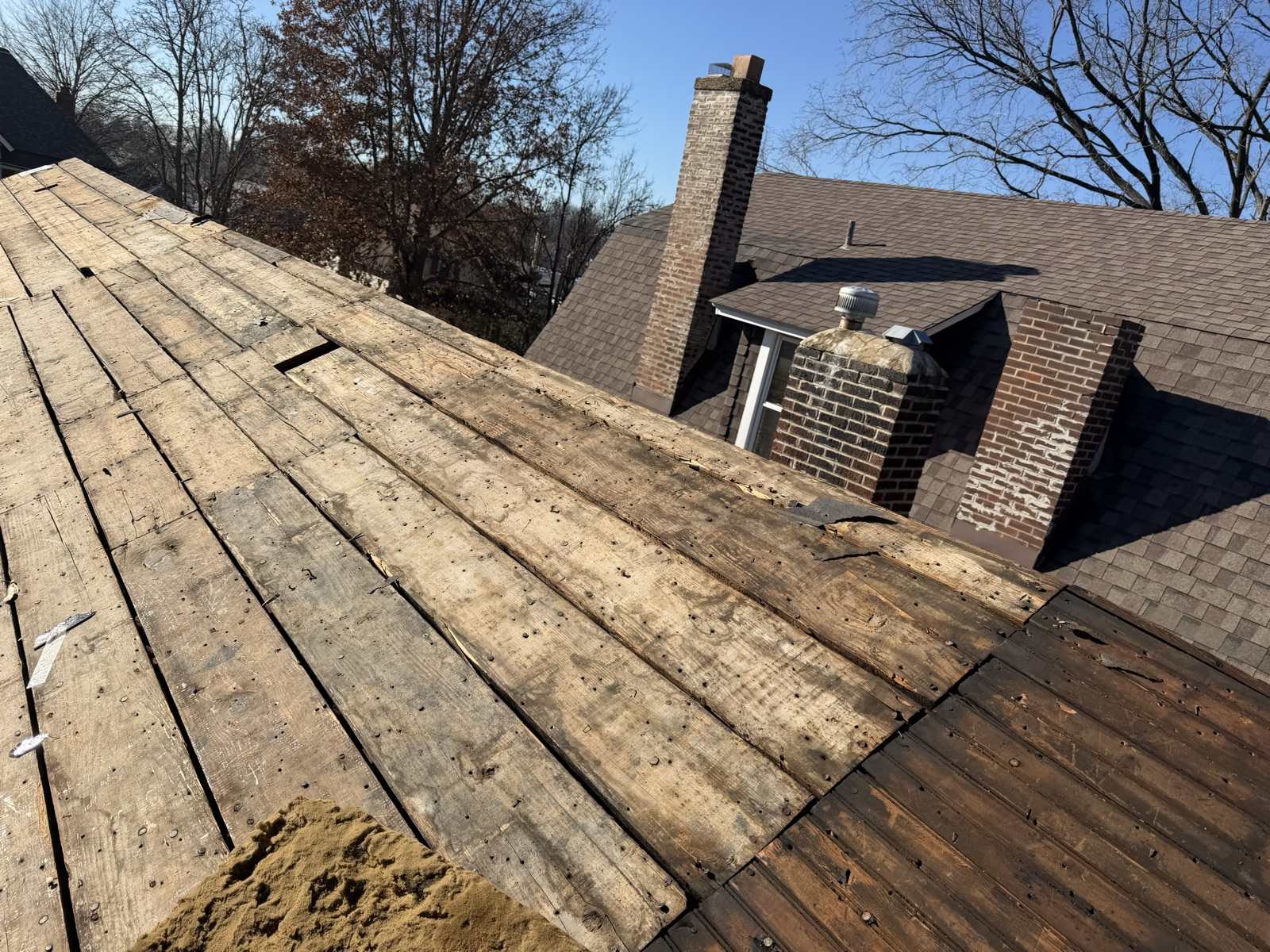 A roof stripped to bare plank decking in the foreground, the boards weathered grey and split around old nail holes, with the adjoining wing of the same house still covered in brown shingles. Two brick chimneys rise behind, one tall and slim and one lower with a stone cap and a metal flue, next to a mushroom shaped attic vent. Bare winter trees and a clear blue sky sit behind.