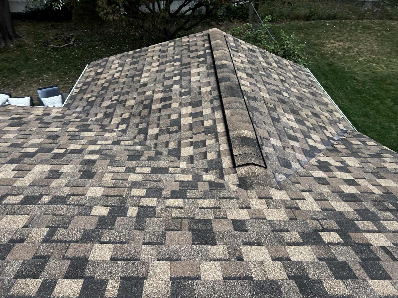 Looking down at the meeting of two hip planes on a roof in multi-tone brown and tan architectural shingles, with ridge cap shingles being run over a black ridge vent strip along the top, and a mown back yard with patio cushions and a shrub line below.
