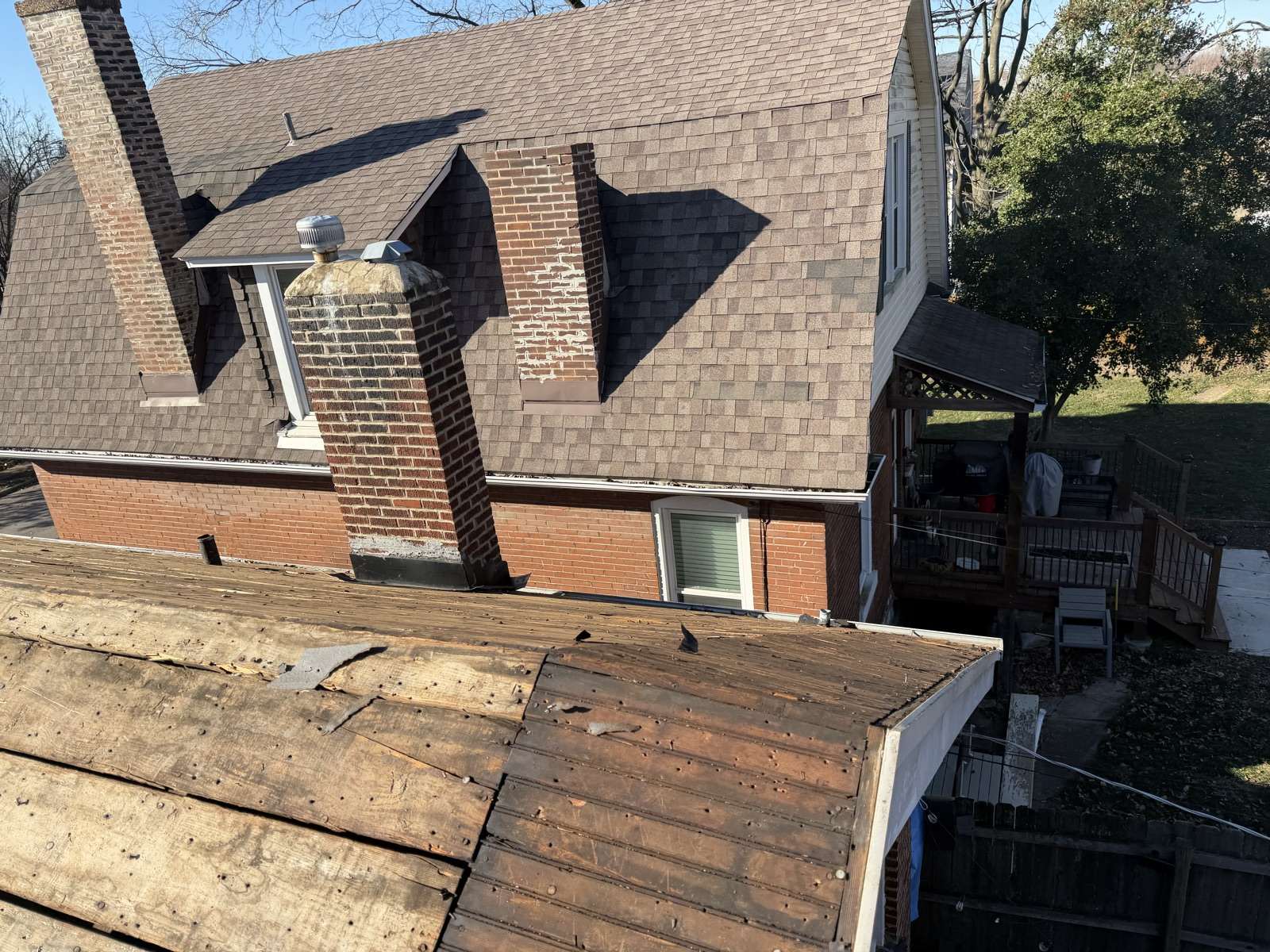A St. Louis brick house mid job. In the foreground the roof of a lower rear wing has been stripped to dark weathered plank decking with torn felt fragments and exposed nails, while the main house behind still carries its old brown shingles. Two brick chimneys rise through it, one slim and one with a stone cap, the brick face patched and spalling, and the eave gutter is packed with leaf debris. A wooden deck, a covered grill and a fenced back yard sit below.