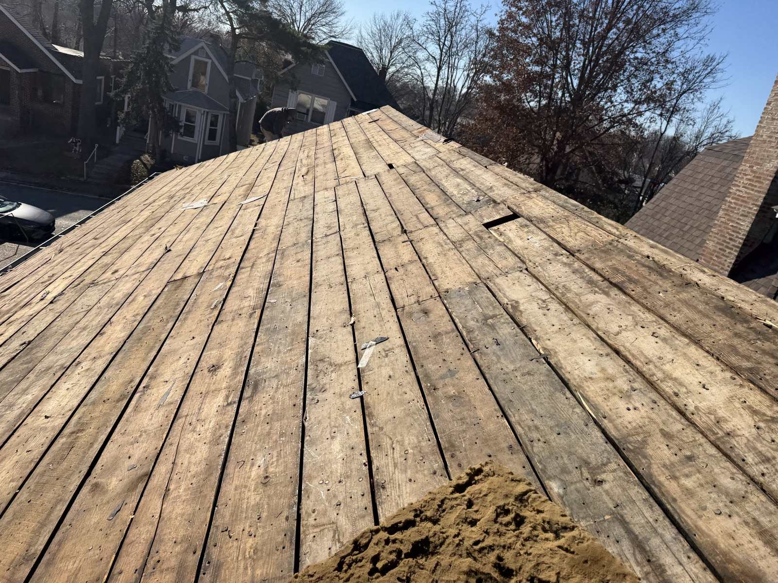 Looking straight up the slope of a roof stripped to bare plank decking, wide sawn boards laid with gaps between them, studded with old nails and scraps of felt, one board split away near the ridge. A pile of sawdust sits in the foreground, a figure is bent over at ground level beyond the far ridge beside a neighbouring grey house, and an older St. Louis street of brick and frame houses with bare winter trees lies below.
