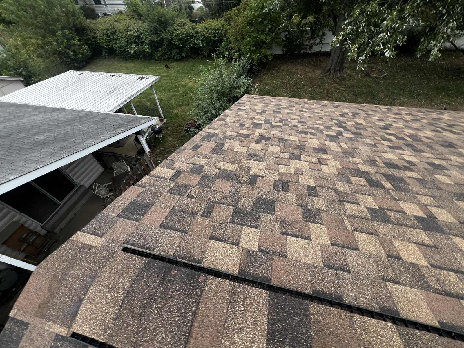 A finished plane of multi-tone architectural shingles seen from above, laid in even courses with a black ridge vent along the near ridge, an older metal patio cover and a shingled porch roof at the left and a green back yard beyond.