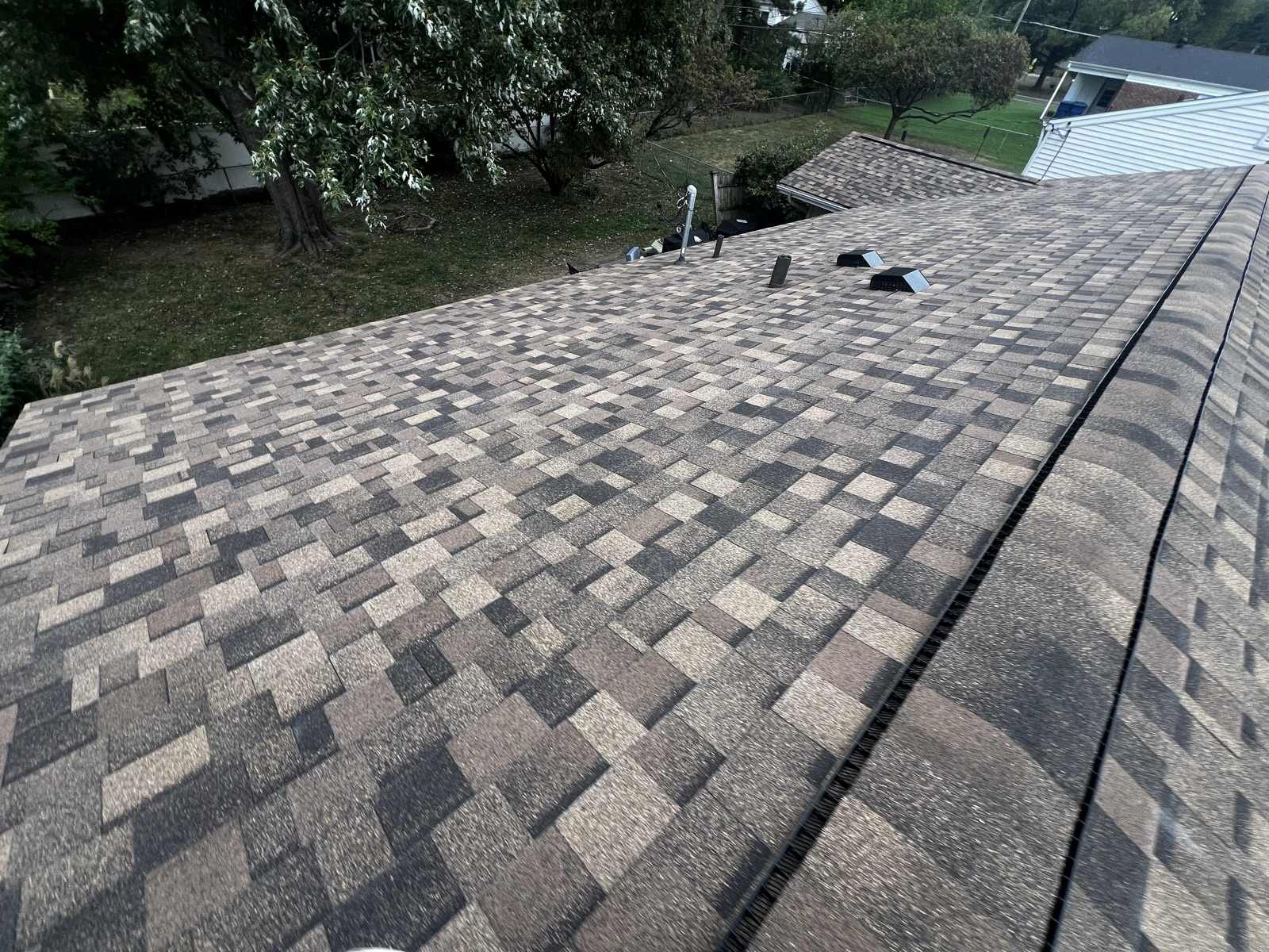 A finished roof in multi-tone brown, tan and charcoal architectural shingles seen along the slope, with ridge cap running over a black ridge vent at the right, three low black box vents and a metal pipe clustered near the far end of the ridge, and a back yard with a mature tree, a chain link fence and a neighbouring white sided house beyond.